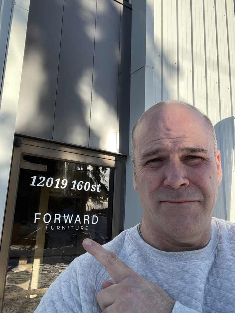 Colin Draper, the Furniture Fella, pointing at the entrance of Forward Furniture Edmonton located at 12019 160 Street NW.