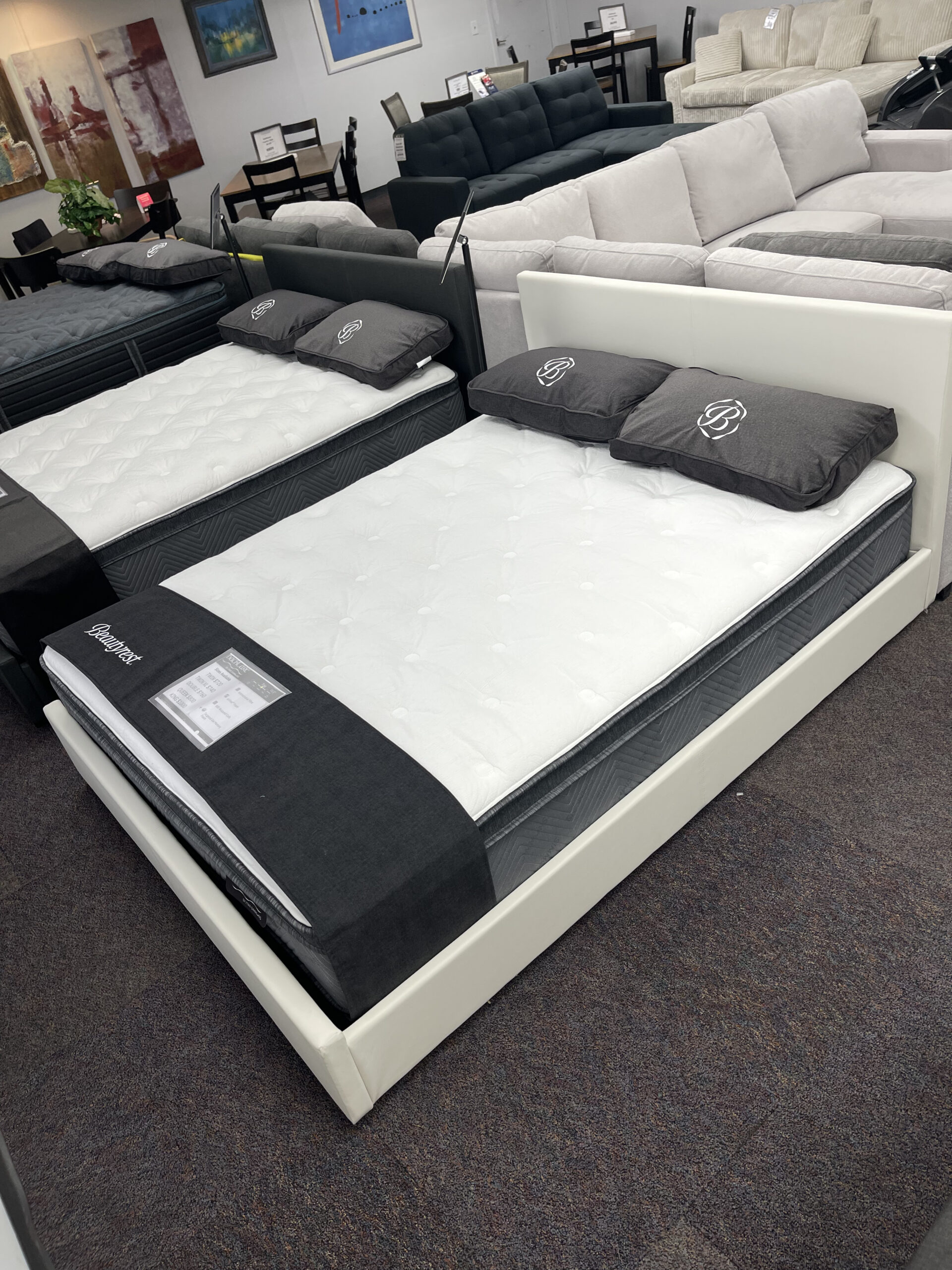 Two Simmons Beautyrest mattresses displayed on white and black bed frames in the Forward Furniture showroom. The beds feature thick white quilted tops, grey branded pillows, and black foot protectors, with sofas and dining sets visible in the background. Simmons Backcare trio at Forward Furniture Edmonton.