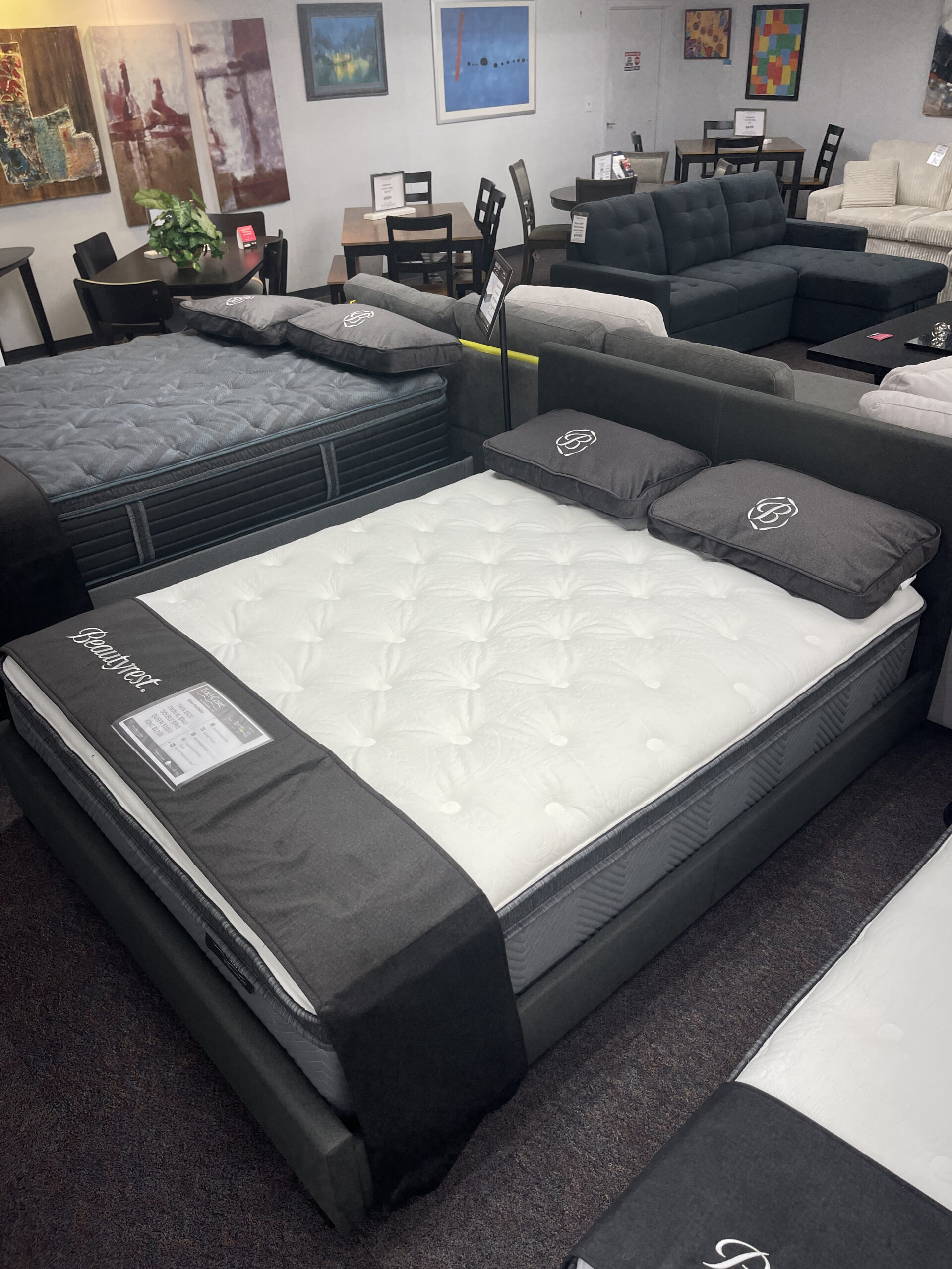 Two Simmons Beautyrest mattresses displayed on white and black bed frames in the Forward Furniture showroom. The beds feature thick white quilted tops, grey branded pillows, and black foot protectors, with sofas and dining sets visible in the background. Simmons Backcare quartet at Forward Furniture Edmonton.