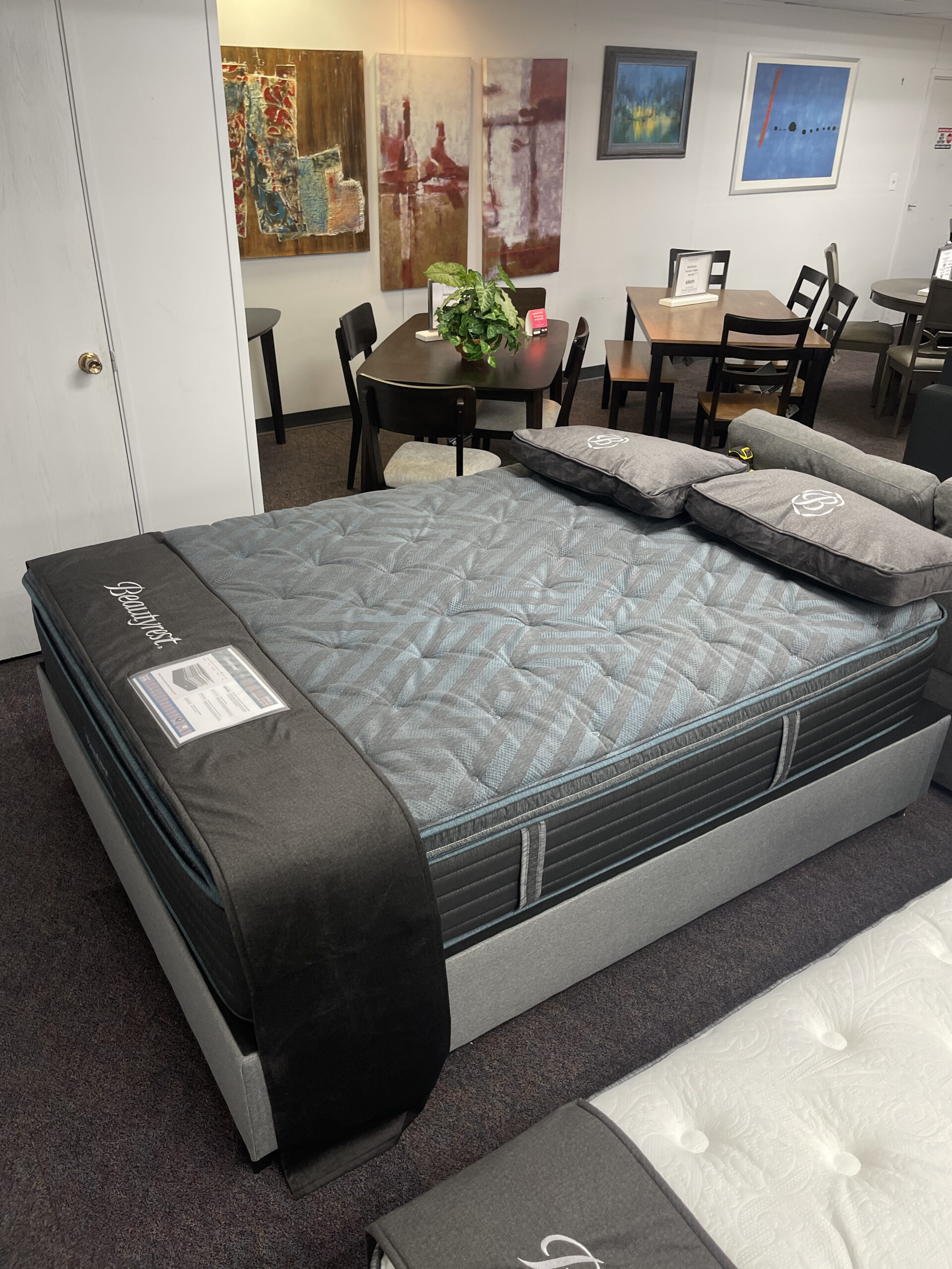 Two Simmons Beautyrest mattresses displayed on white and black bed frames in the Forward Furniture showroom. The beds feature thick white quilted tops, grey branded pillows, and black foot protectors, with sofas and dining sets visible in the background. Beautyrest Erato at Forward Furniture Edmonton.