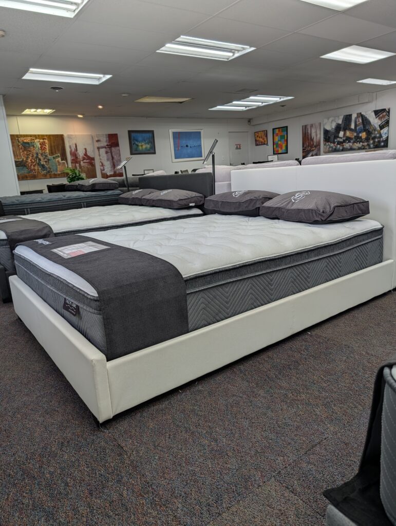 Simmons Beautyrest mattresses displayed on white and black bed frames in the Forward Furniture showroom. The beds feature thick white quilted tops, grey branded pillows, and black foot protectors, with sofas and dining sets visible in the background. Simmons Backcare trio at Forward Furniture Edmonton.