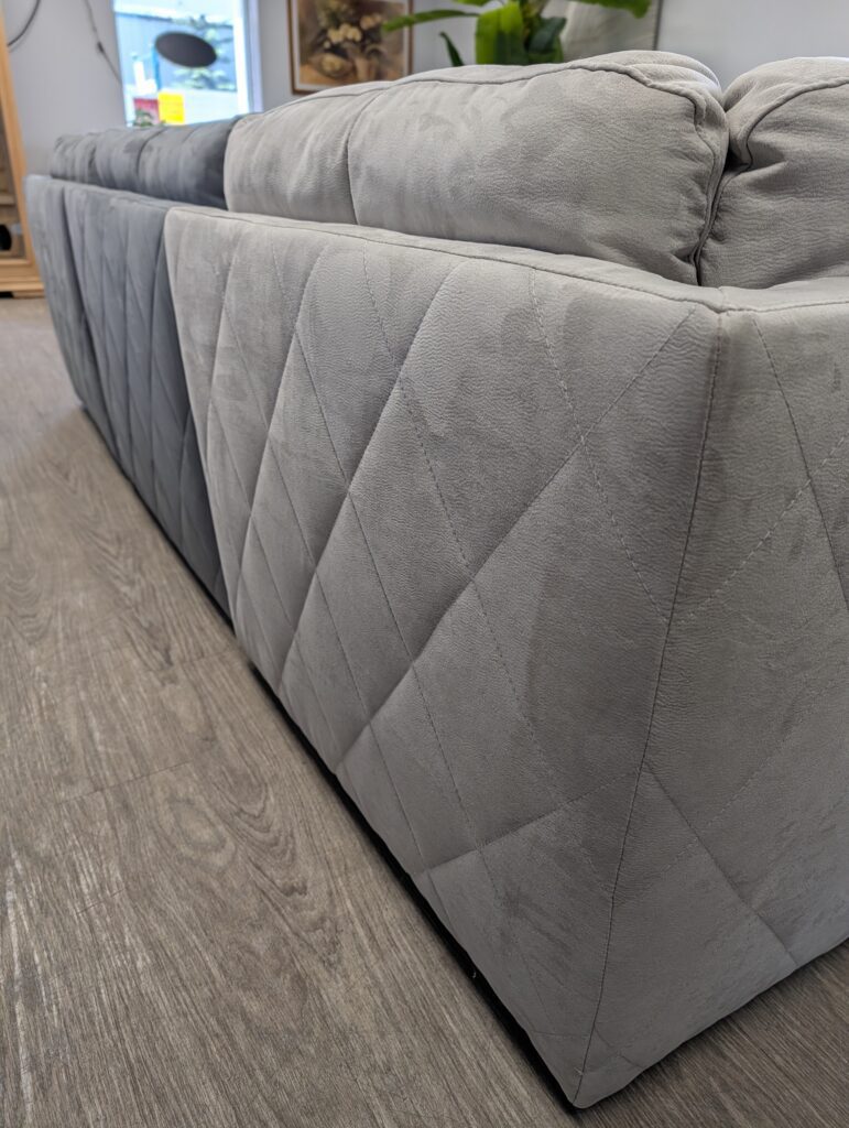 Close-up of a grey fabric sofa featuring diamond-quilted stitching on the back and side, resting on a wood-grain floor.