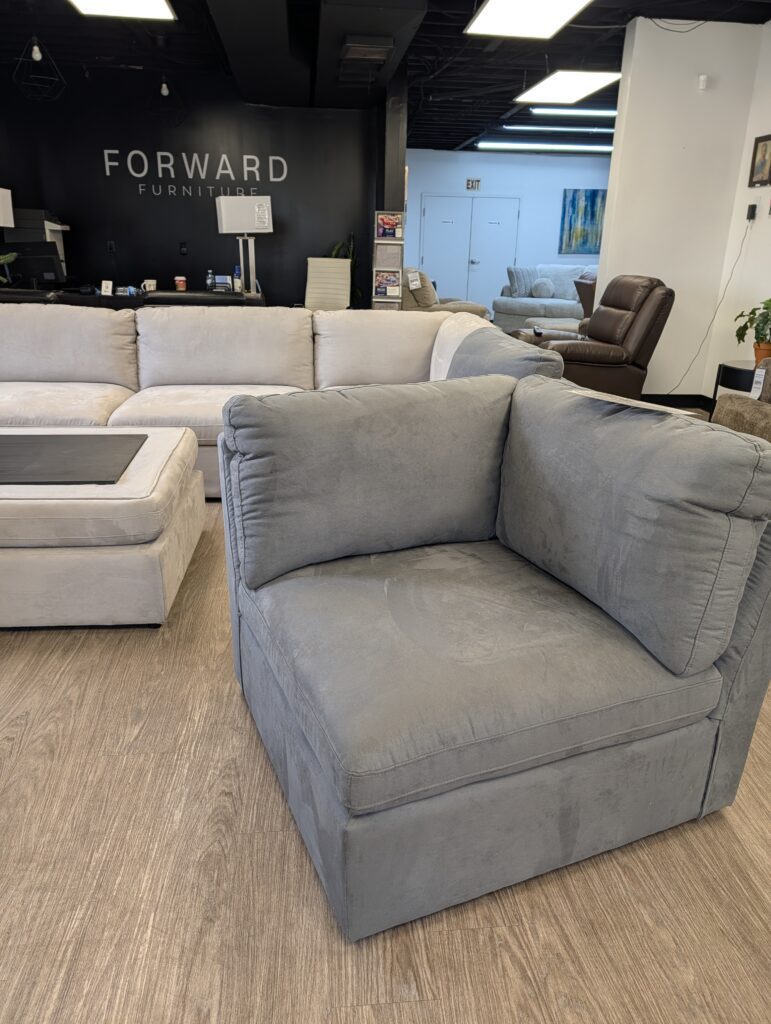Grey fabric armless sofa piece in a furniture showroom with wood-grain floors. A white sectional, brown leather recliner, and a black wall featuring a "FORWARD FURNITURE" sign are in the background.